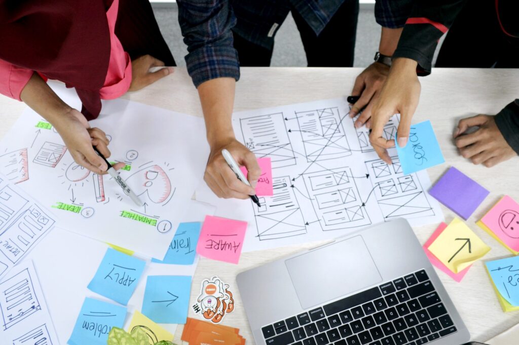 peoples' hands putting post its on paper and working in front of a laptop